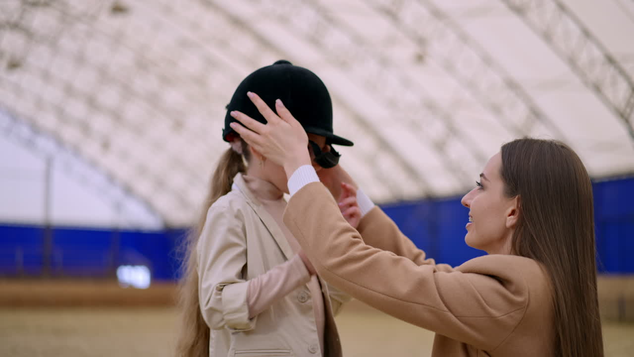 Long-haired woman in a beige coat kneeled near a young girl. Lady puts jockey hat on daughter's head but it's little too big.