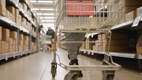low ground view shows grocery shopper wearing boots rolling trolley down lit supermarket aisle, boxes lining shelves, other customers and carts blurred in distance conveying retail atmosphere