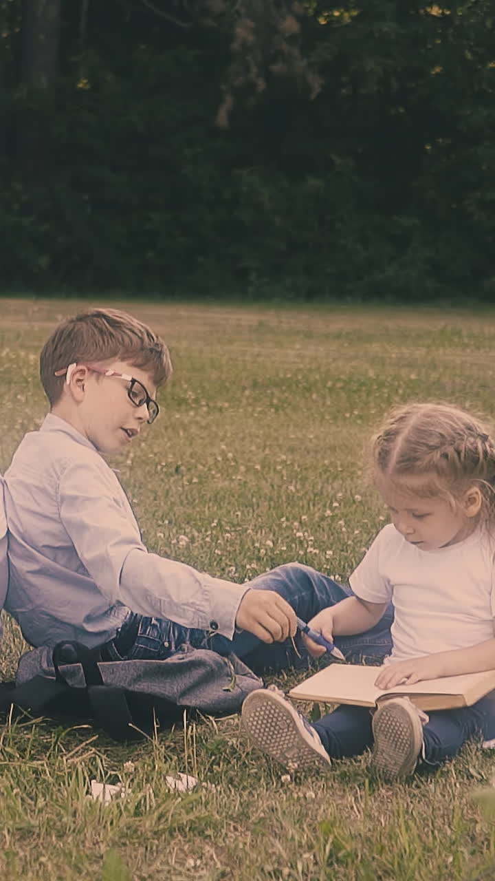 schoolboy eats snack and brother helps junior schoolgirl with writing in copybook sitting in school garden slow motion