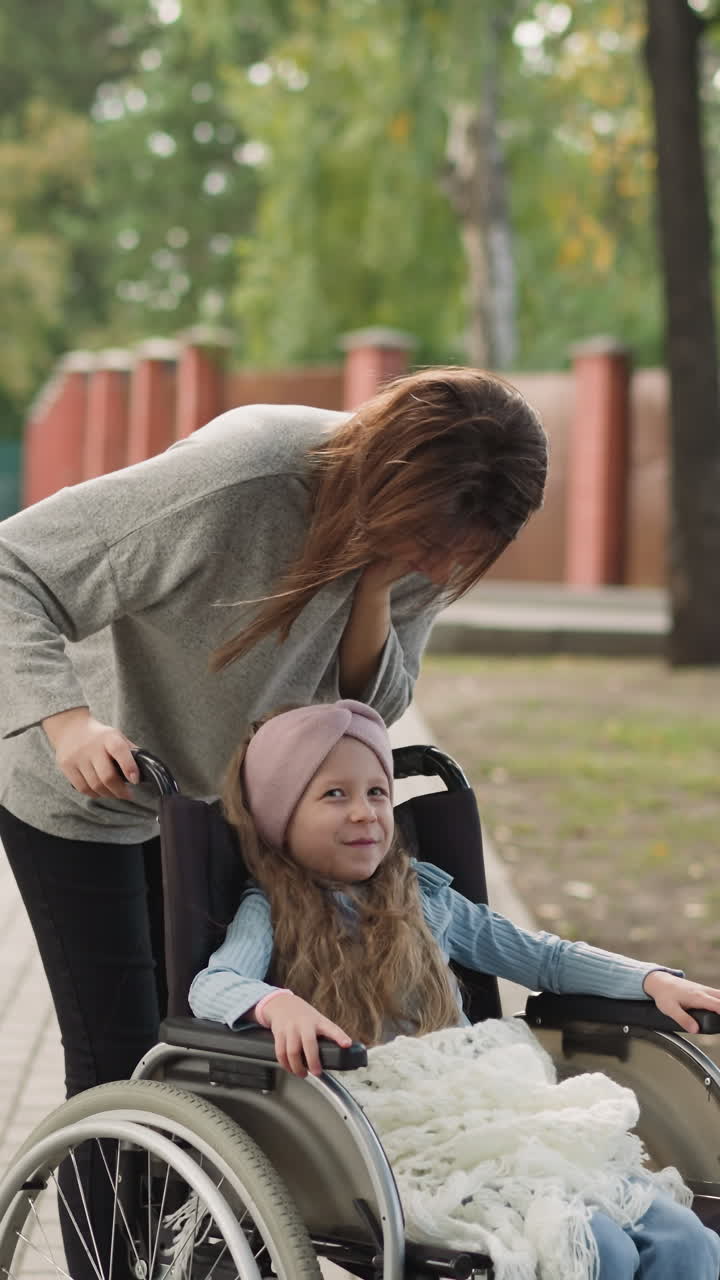 madre feliz besa a su hija sentada en silla de ruedas en el pavimento de la calle. mujer pelirroja disfruta de caminar en el parque con una niña con una condición de salud crónica