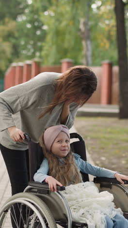 happy mother kisses daughter sitting in wheelchair on street pavement. redhead woman enjoys walking in park with little girl with chronic health condition