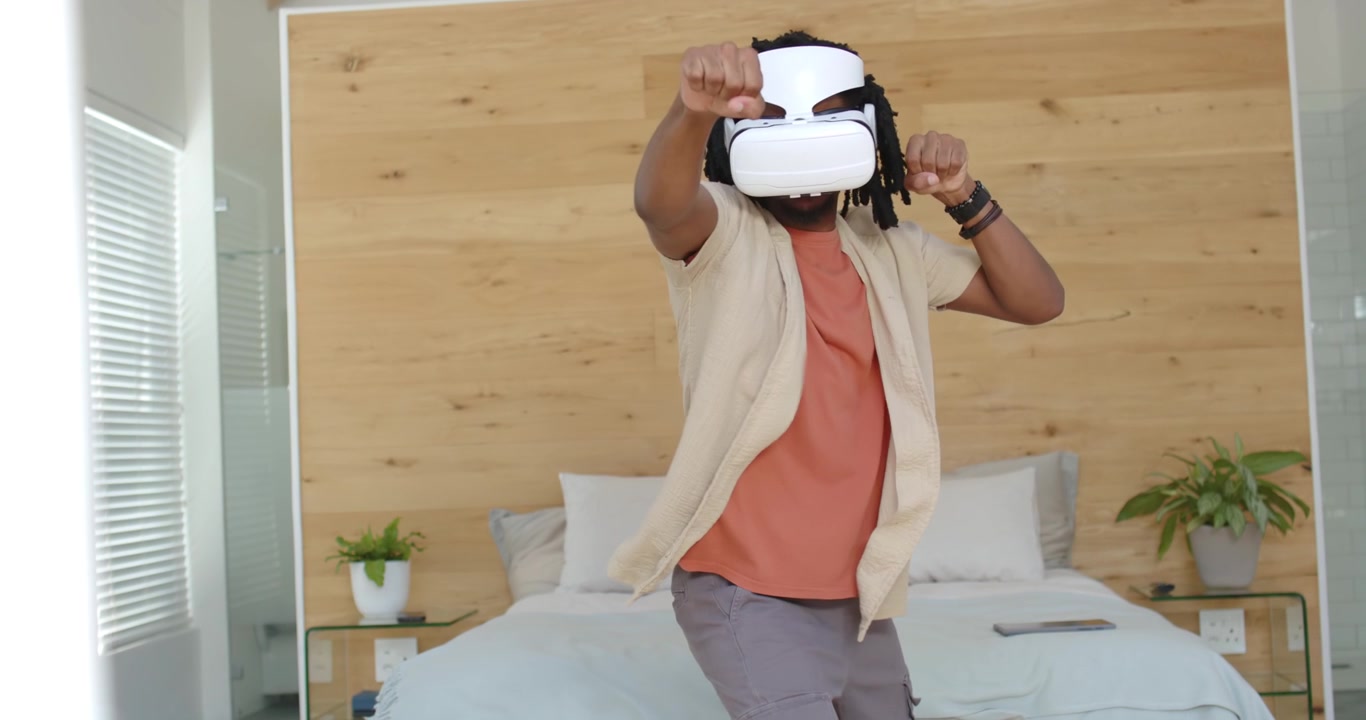 Uomo afroamericano che indossa un visore VR bianco in camera da letto, controllando il visore e sferrando pugni per il fitness.