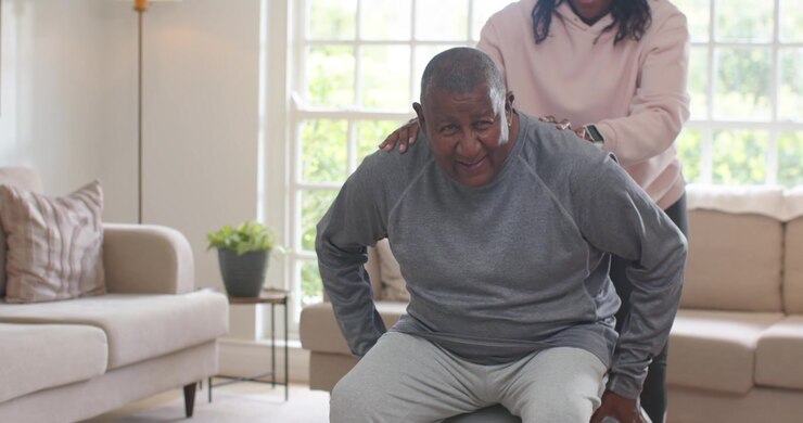 African American woman gesturing, guiding mature man onto stability ball for balance at home