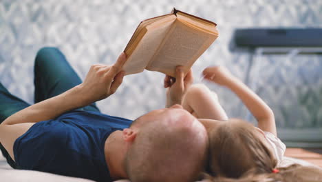 father reads book to little daughter doing homework on floor