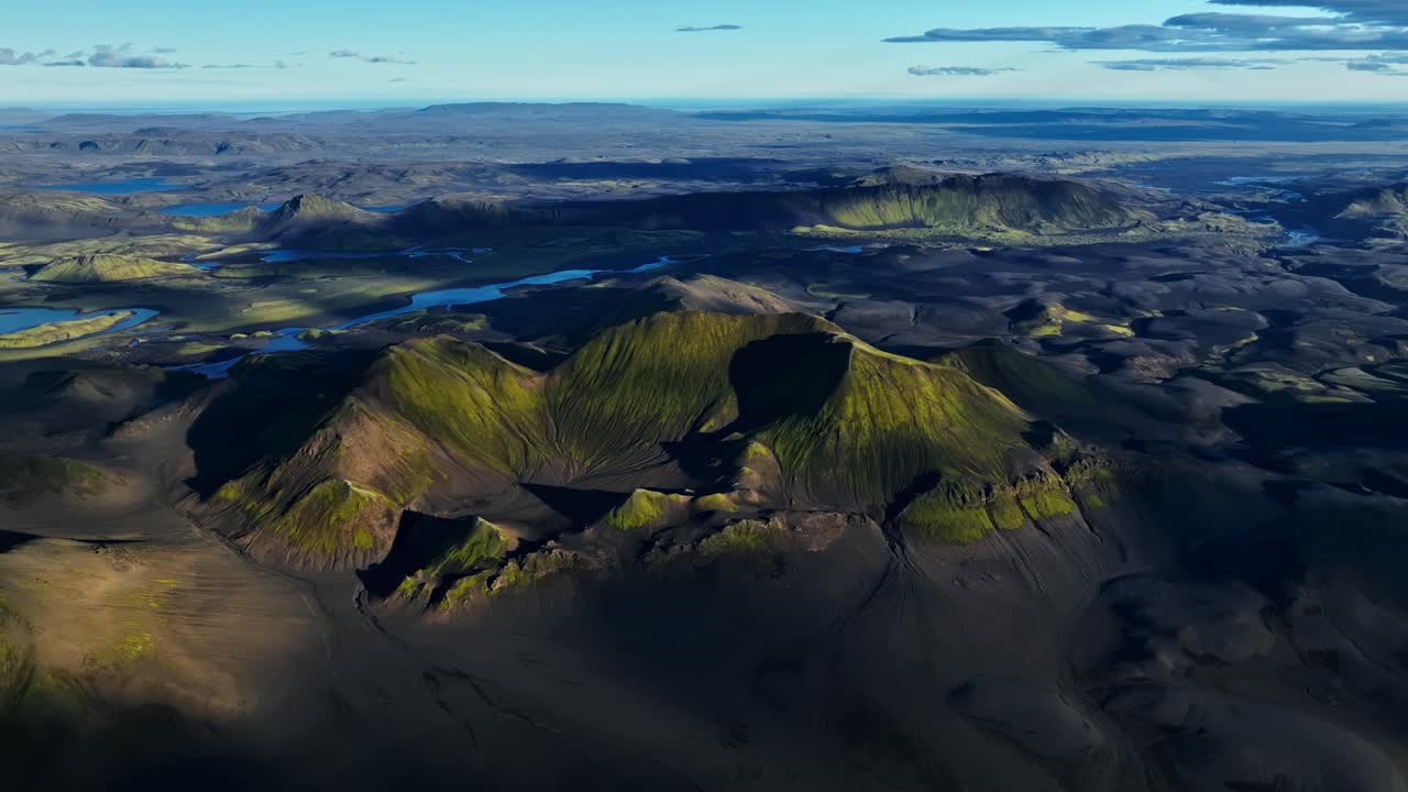 Drone view over Iceland’s dark volcanic plains and moss-covered craters, revealing layered ridges, distant lakes and long evening shadows across the highlands