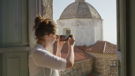 glückliche frau, die ein smartphone benutzt, fotos macht, spaß am teilen von urlaubserlebnissen macht, schöne aussicht auf dem balkon fotografiert, sonnenbrille trägt