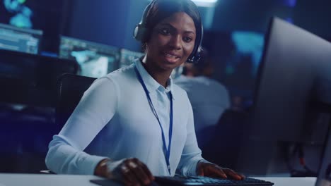 joyful beautiful technical customer support specialist is talking on a headset while working on a computer in a call center control room filled with colleagues, display screens and data servers.