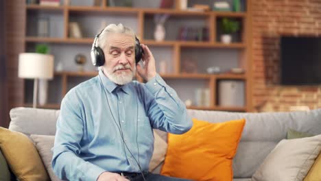 cheerful senior retirement gray-haired man listening to music on headphones while sitting at home on the couch