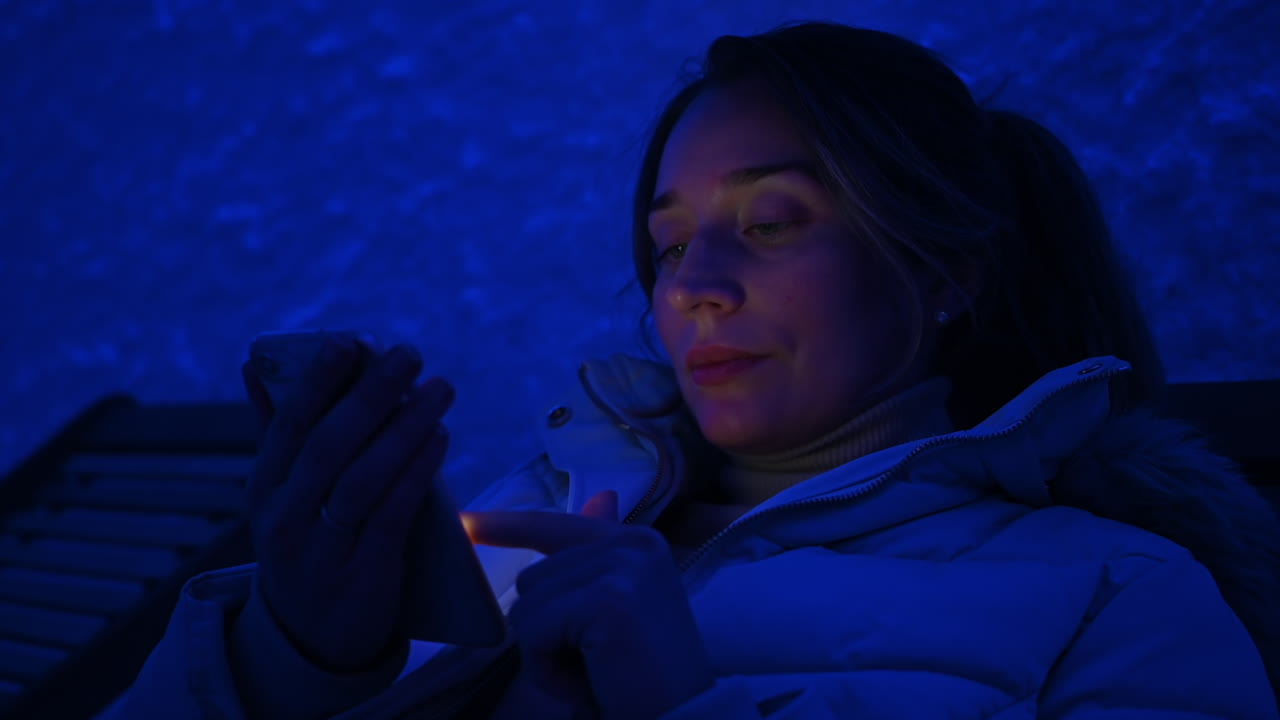 Woman relaxing on a wooden chair in a salt room with blue light, working on mobile phone