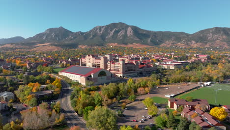 aerial moving forward view of cu boulder campus and stadium surrounded by green and yellow fall trees with the rocky flatiron mountains in the background in the front range of colorado