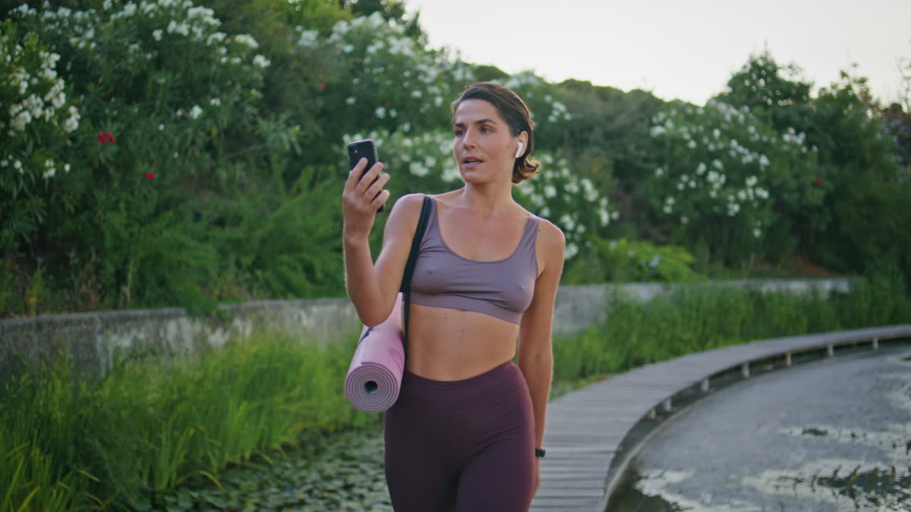mujer deportiva sonriente llamando por teléfono móvil paseando por el callejón del parque en primer plano