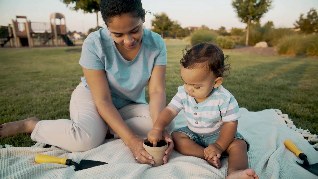 Placing pot mother blue-top steadying pot at park, toddler striped-tee scooping soil learning
