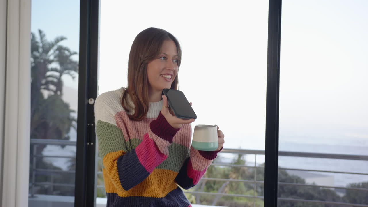 Holding smartphone and mug, woman smiling and talking in modern home setting