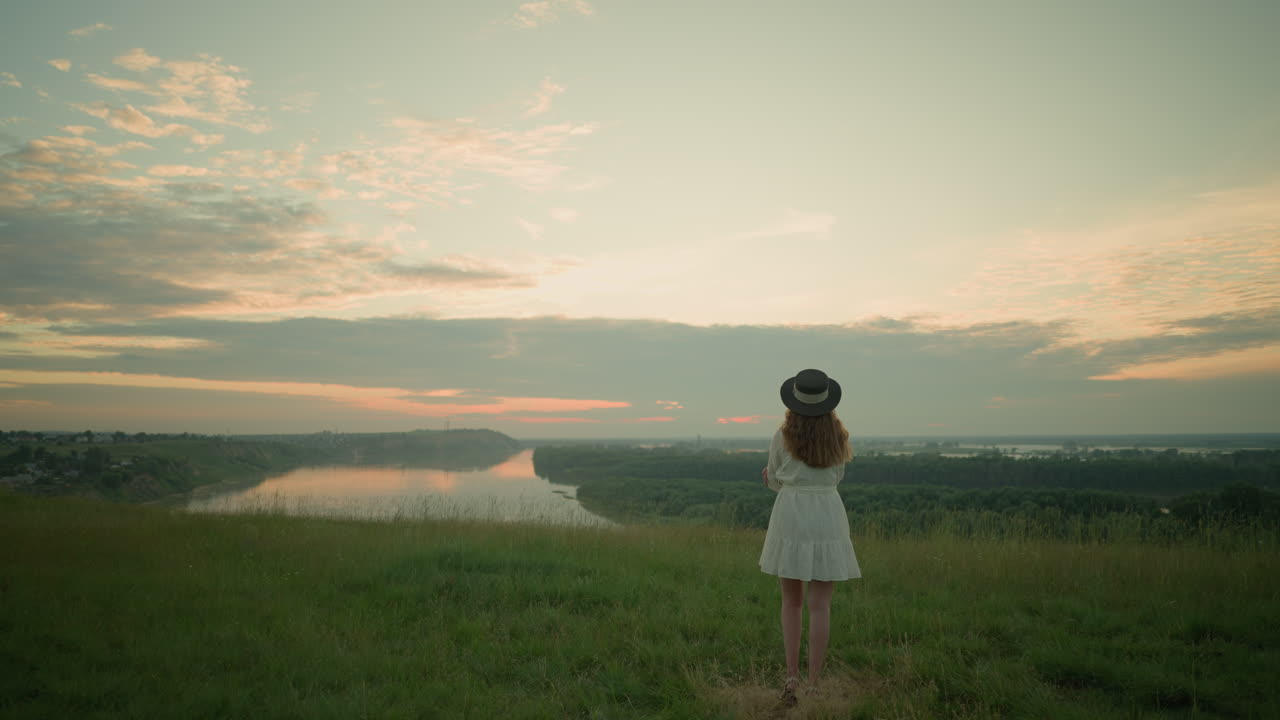A woman in a white dress and hat stands with folded arms, thoughtfully in a grassy field overlooking a tranquil lake at sunset. She gazes at the peaceful environment