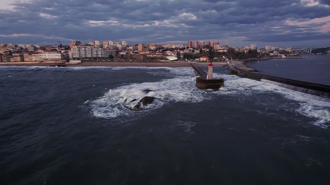 Drone push in sweeps in toward the Felgueiras Lighthouse and surf-lashed breakwater at Foz do Douro, catching Porto’s oceanfront skyline as the sun sets over the Atlantic