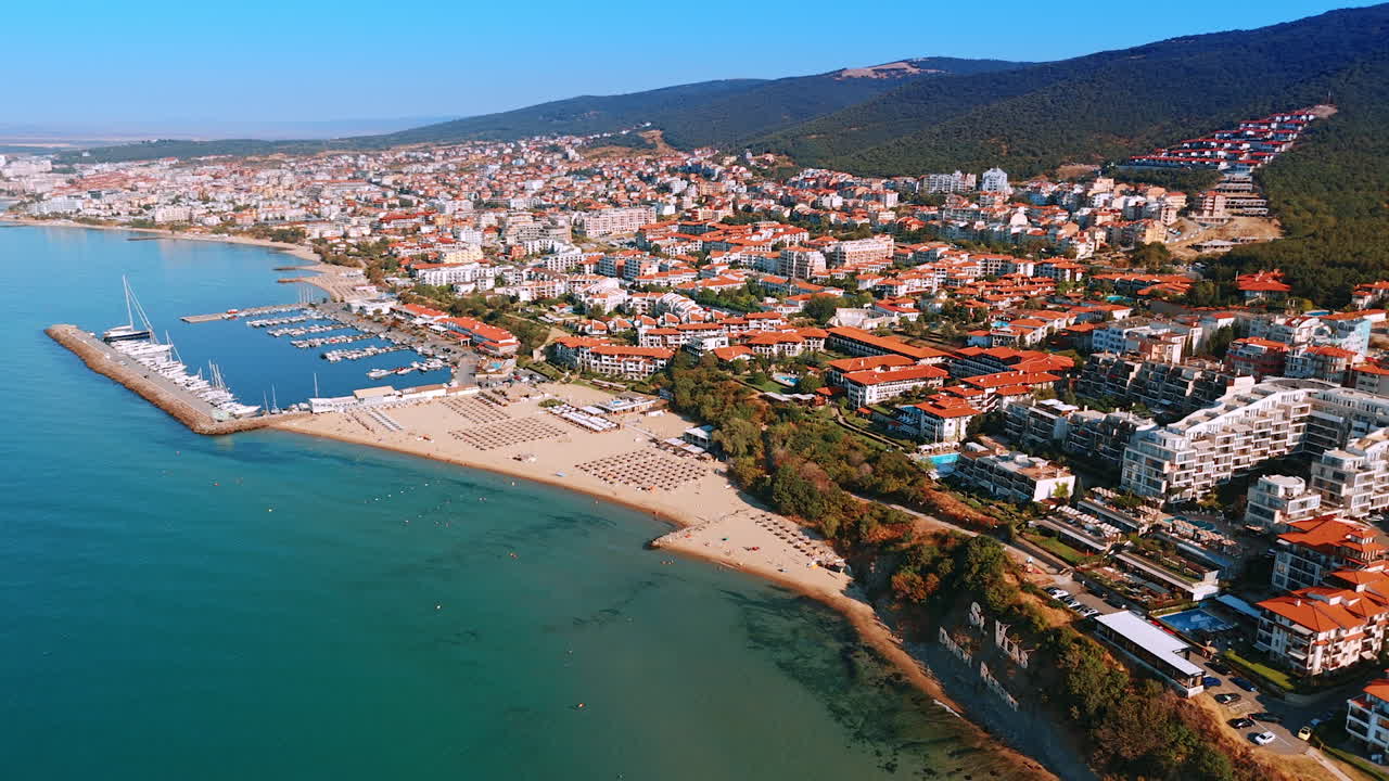 Beach town by the marina. The vibrant coastal town features white buildings, a busy marina, and a sandy beach under clear blue skies