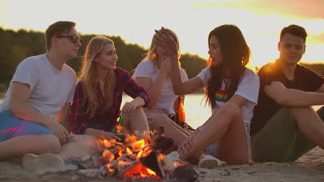 two boys and three girls are sitting around bonfire on the beach with beer. they are talking to each other at sunset and enjoying the summer evening on the river coast.