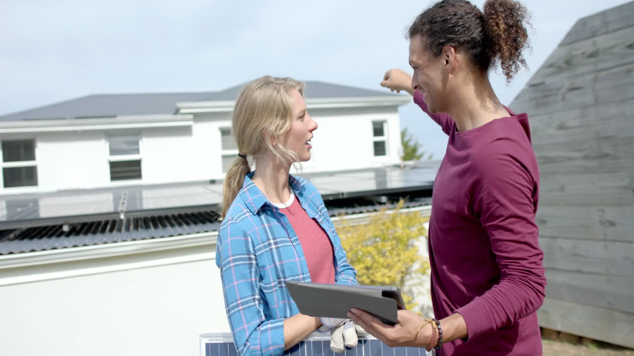 Busy diverse couple using tablet and holding solar panel in garden, slow motion