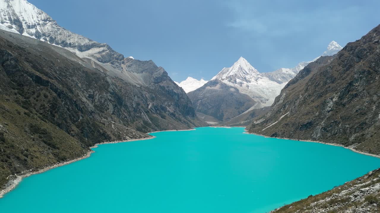 A spectacular aerial shot pans alongside a steep mountain, revealing the vast, vibrant turquoise waters of Laguna Parón and the snow-capped peaks of the Cordillera Blanca in Peru
