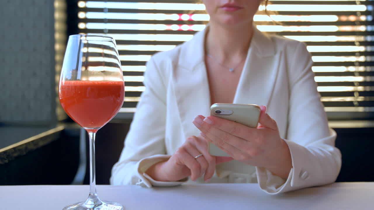 Woman in white blazer scrolling on the phone with a carrot juice on the table at a restaurant