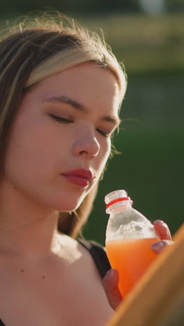 mujer sentada al aire libre sosteniendo un libro en una mano mientras toma una botella de jugo en la otra, continuando a leer, el fondo borroso presenta un poste de lámpara y una colina verde
