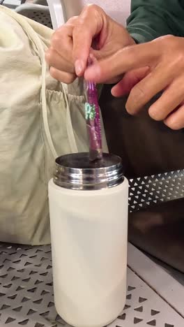 person mixes a beverage in a thermos while seated.