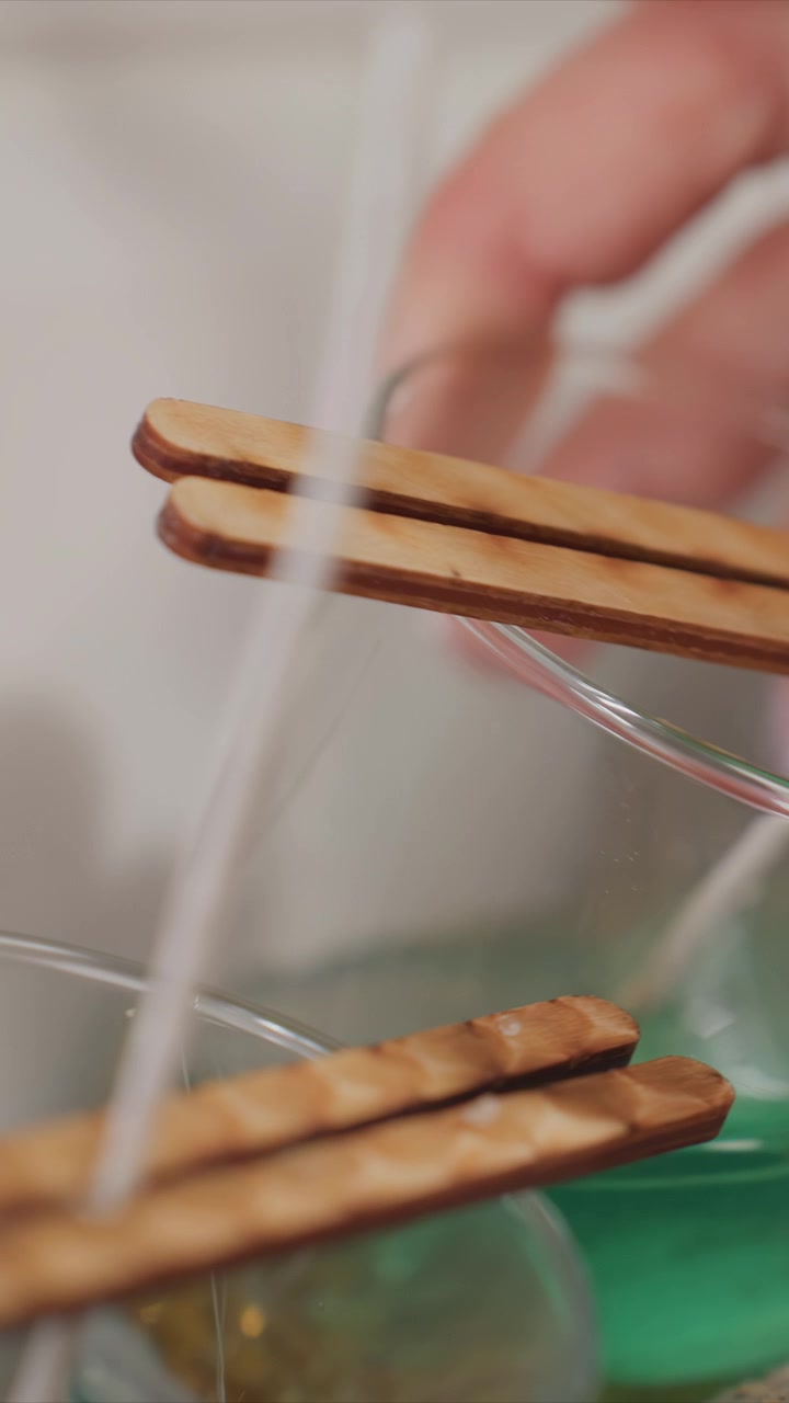 Primo piano di una mano con unghie curate che immerge stoppini di legno in cera liquida verde per la fabbricazione di candele. Il processo prevede il posizionamento attento dei bastoncini per garantire una copertura uniforme, evidenziando.