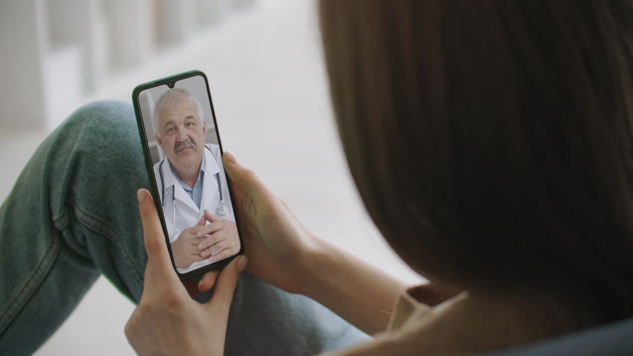 Female medical assistant wears white coat headset video calling distant patient on smartphone. Doctor talking to client using virtual chat telephone app. Telemedicine remote healthcare services