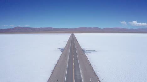 Car-drives-on-straight-empty-road-through-Argentine-salt-flats,-aerial