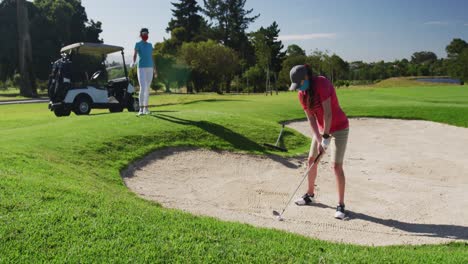 Two-caucasian-women-playing-golf-wearing-face-masks-one-taking-shot-from-bunker