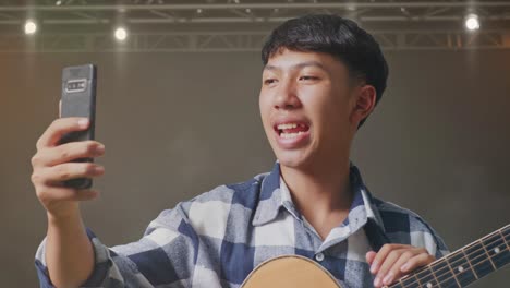 primer plano de un adolescente asiático con la mano agitando la guitarra y haciendo una llamada de video en el teléfono inteligente mientras está de pie en el escenario