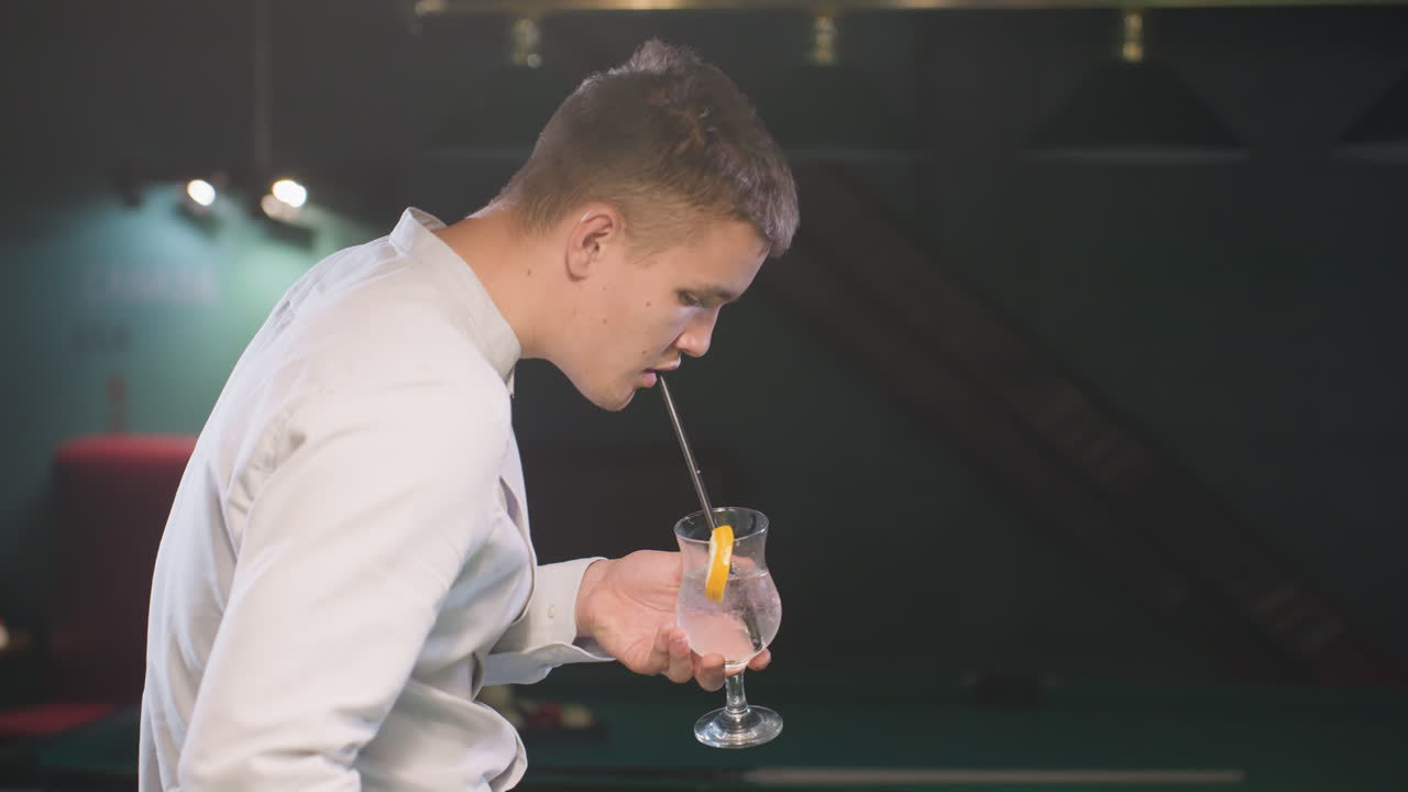 셔츠를 입은 젊은 남자가 술을 마시면서 검은 을 가진 레몬 음료 한 잔을 들고 있는 클로즈업. 비언트 조명은 편안한 분위기를 강조하여 현대적이고 세련된 환경을 만니다.