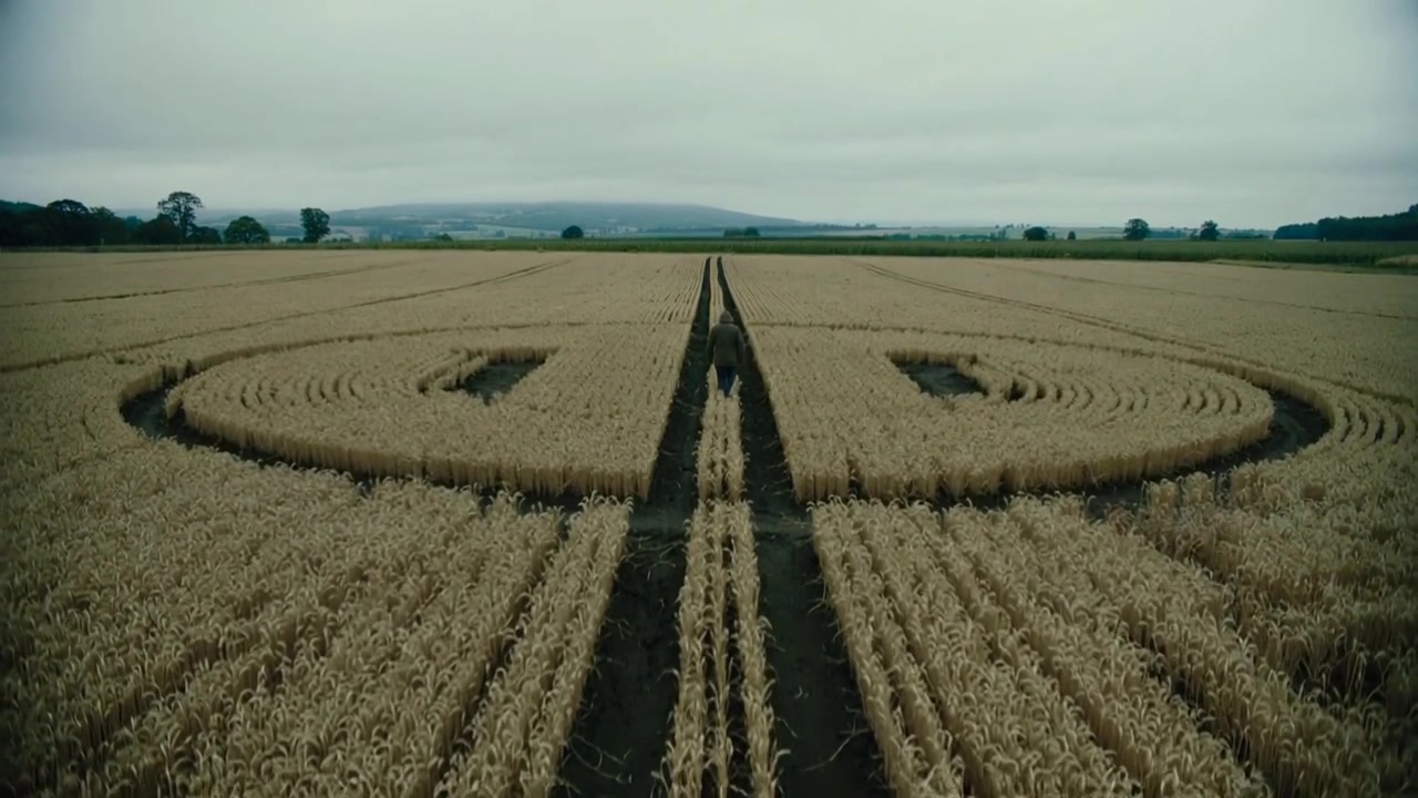 Walking figure in dark coat moving along tracks to horizon, camera pulling back showing crop circle