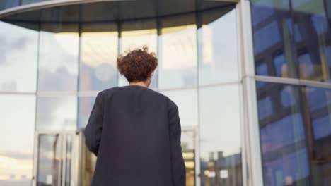 young man in eyeglasses texting on cell phone, looking around and entering modern glass office building with revolving door, briefcase in his hand