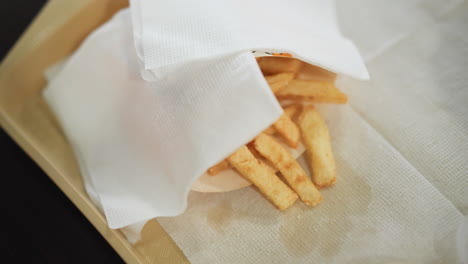 primer plano de papas fritas doradas crujientes parcialmente cubiertas con una servilleta en una bandeja, con una mano alcanzando las papas fritas, la escena captura un momento de cena relajado y casual