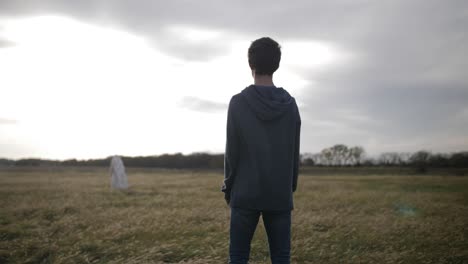 a young teenage boy stands in a field and looks at jesus christ - or man wearing a white robe - in the distance with bright sunlight shining