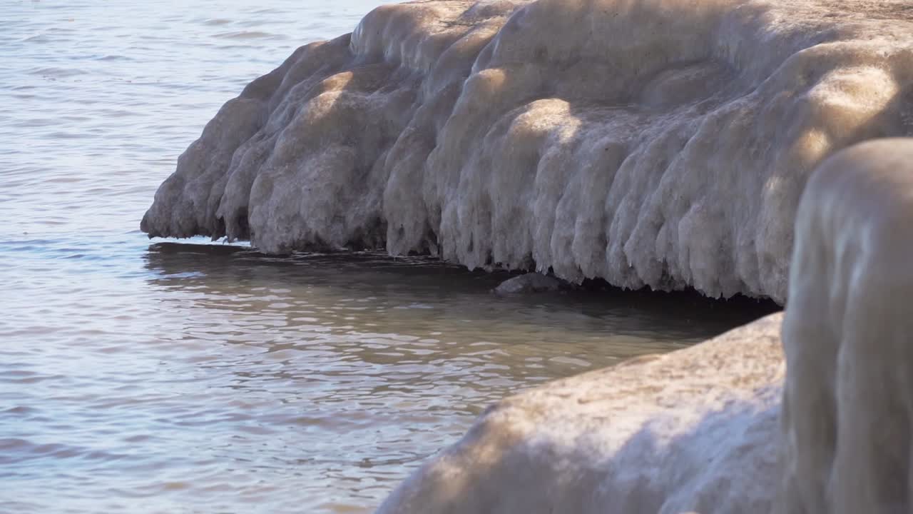 lake water waves flow under over hangin ice formation, water reflects blue sky.