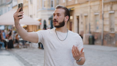 young man blogger taking selfie on smartphone video call online with subscribers in city street