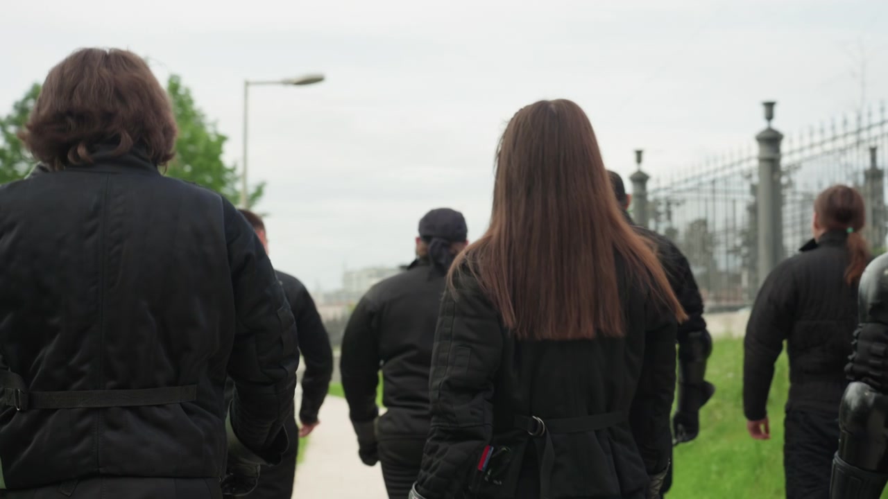 Gruppo di persone con abbigliamento imbottito nero che camminano all'aperto vicino a una recinzione metallica e vegetazione. Le persone indossano equipaggiamento protettivo, inclusi guanti, copricapi o bandane, con alcuni che trasportano attrezzatura.