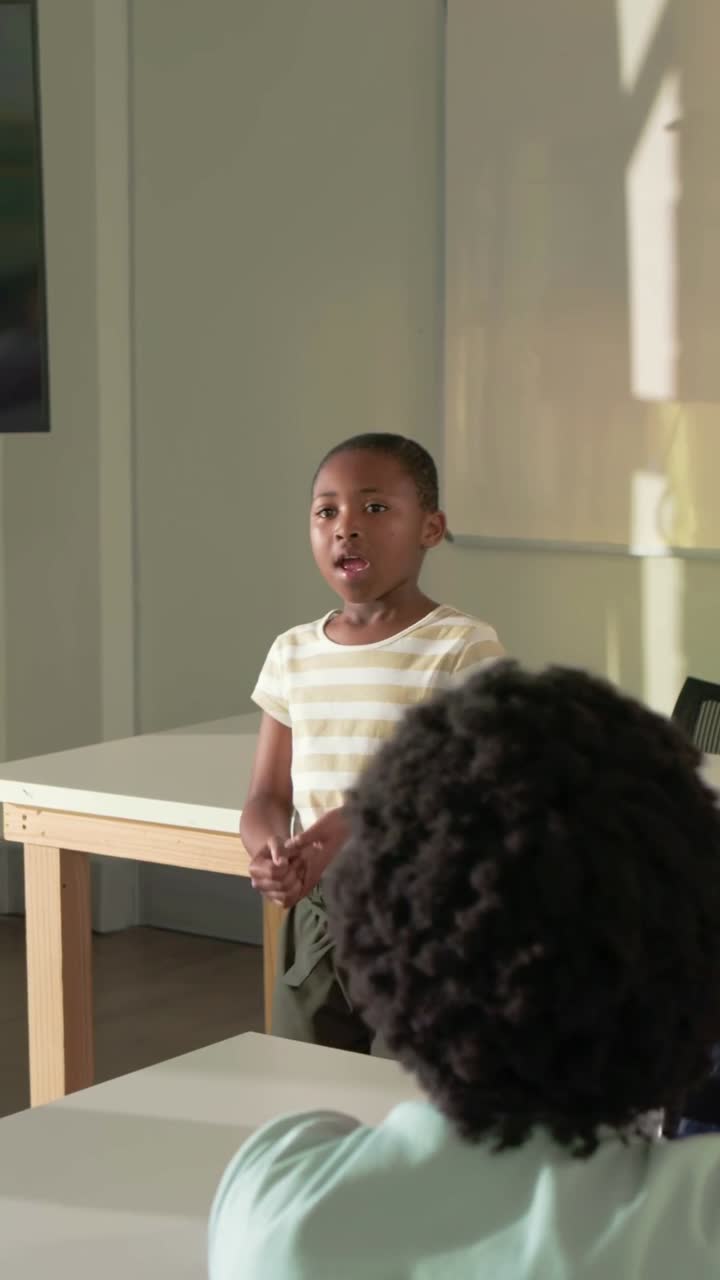 Vertical video: Starting talk, pupil at table, speaking to peers by board wearing striped tee
