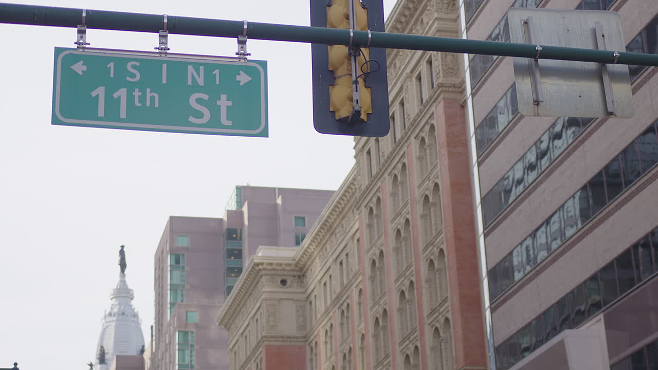 Slow-motion clip of the 11th St sign gently swaying in the wind on a traffic light, with Philadelphia’s Capitol dome, the Ben Franklin statue, and nearby buildings visible in the scenic background.