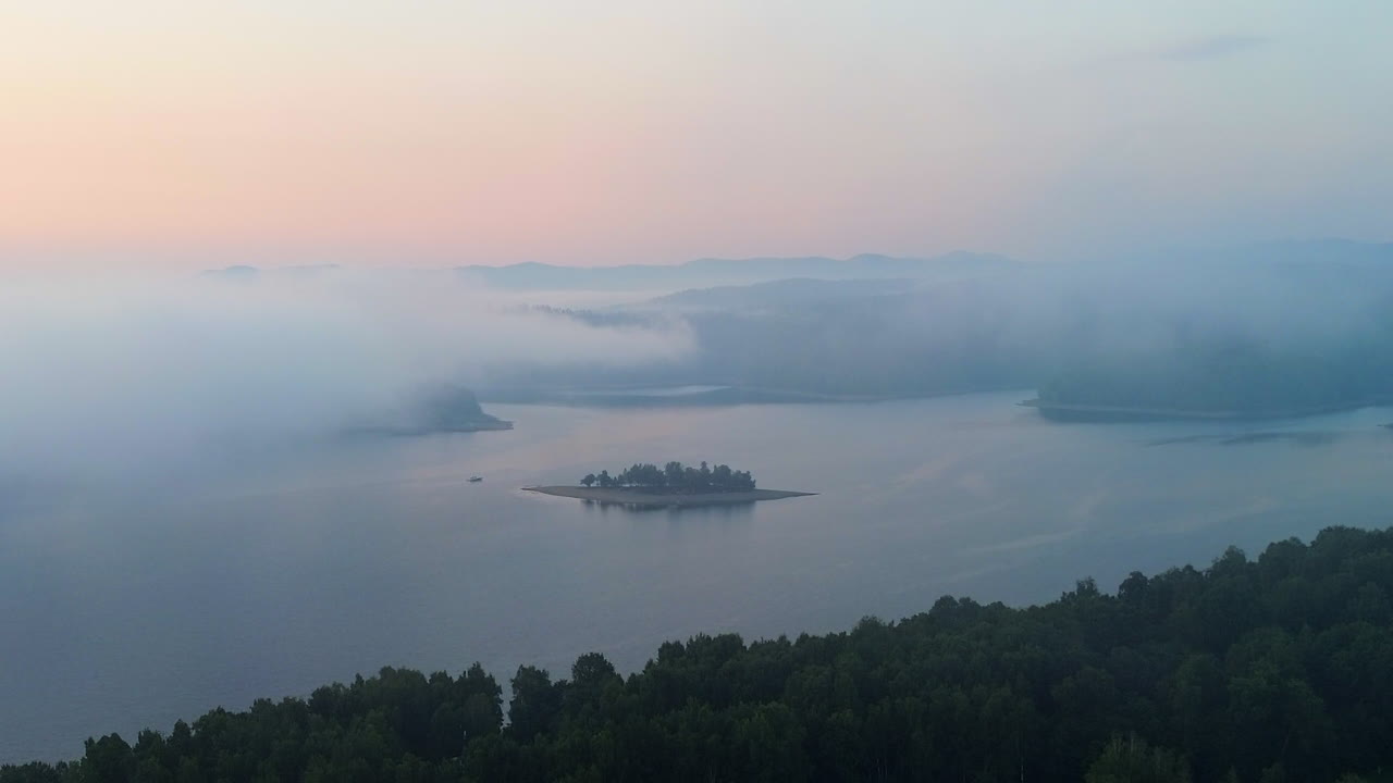 Aerial shot of small forested island surrounded by mist over a calm lake at dawn with soft pastel sky tones. Distant mountain silhouettes. Solina, Poland