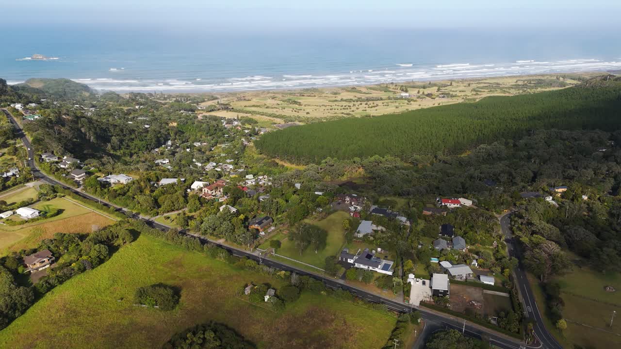 Green landscape with fields and main street leading to Muriwai Beach with Ocean in Background. Aerial tilt up wide shot. Muriwai Town with beach at west coast of Auckland, New Zealand.