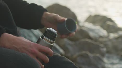 el hombre termina de beber, cierra el termo y se va mientras las olas del mar lavan las rocas en el fondo durante la hora dorada