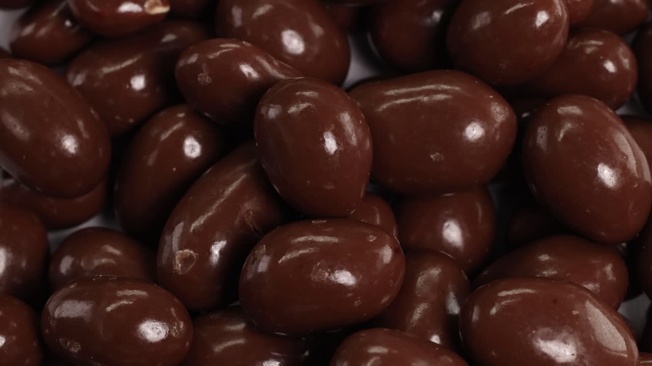 Chocolate-covered peanuts cascade smoothly, creating a rich, textured pattern. Warm lighting enhances the glossy surface, emphasizing movement and indulgence