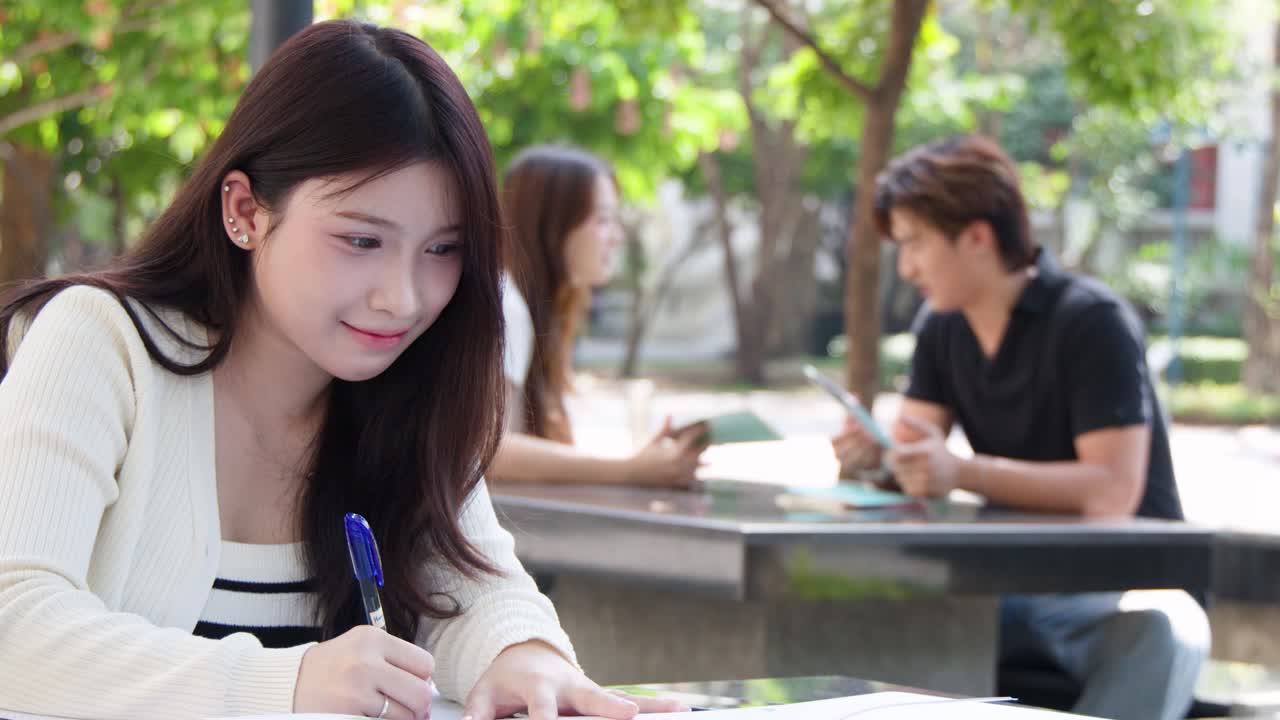 一位泰國學生坐在戶外校園的桌子旁，正在她的筆記簿上書寫。