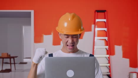 close up de hombre asiático pintor con casco de seguridad mirando a la computadora portátil luego gritando gol celebrando mientras estaba de pie con la pared de pintura en la habitación