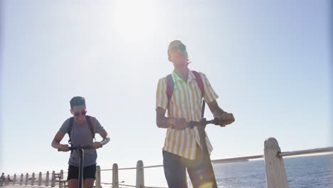 Happy-diverse-gay-male-couple-using-scooters-at-promenade-by-the-sea,-slow-motion