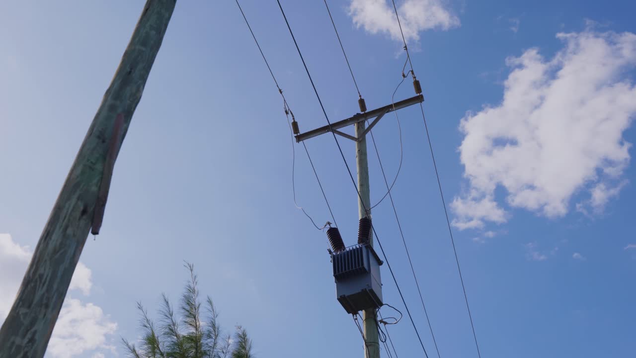 Tall wooden power pole stands against a bright blue sky. Wires stretch across the pole to surrounding greenery, capturing a serene afternoon atmosphere.