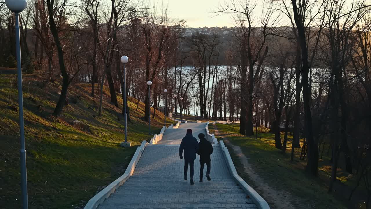 Due giovani uomini alti vestiti pesanti scendono le scale verso il parco Valea Morilor tra lampade e alberi senza foglie. Scatto al tramonto. Vista dall'alto. Chișinău, Moldavia